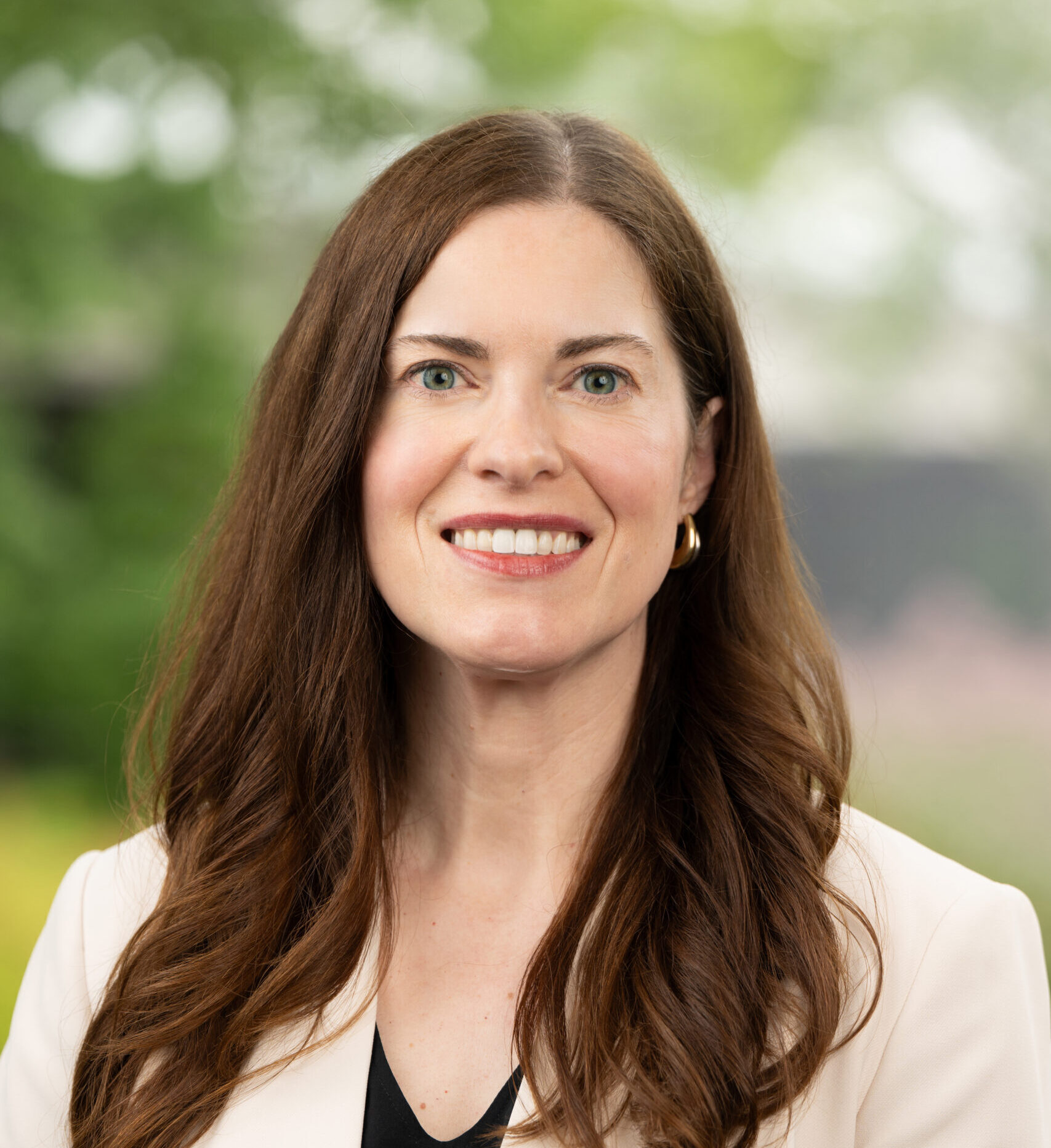 A smiling person with long brown hair, wearing a light-colored blazer over a black top, stands outdoors with a blurred green background.